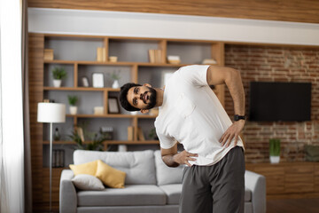 Arabian bearded man dressed in sport clothes holding hands on hips and bending body to sides. Sporty young athlete spending free time for domestic workout.