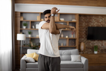 Obraz na płótnie Canvas Handsome focused indian man stretching arms for bodybuilder fitness and muscle warm up at cozy living room. Wellness, sport activity and people concept.