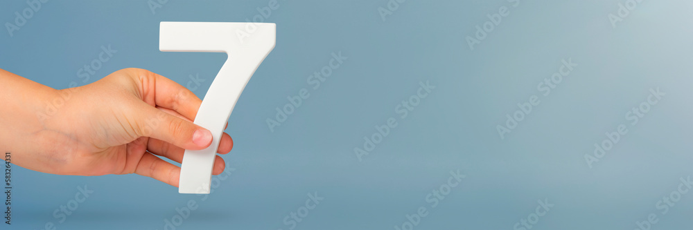 Poster Number seven in hand. Hand holding white number 7 on blue background with copy space. Concept with number seven. Birthday 7 years, percent, seventh grade or day.