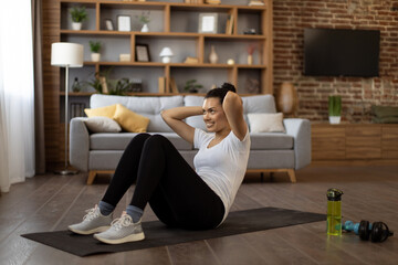 Beautiful fitness woman with dark hair lying on yoga mat and doing abs exercises at living room. Multiracial young lady in activewear having active training alone at home.