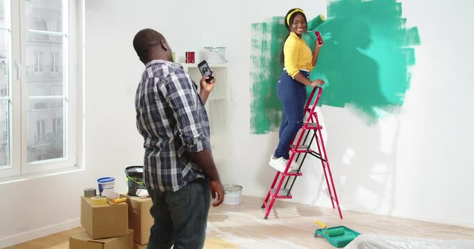 Loving handsome African-American man standing in flat during renovation taking picture of beautiful African woman painting walls with green paint posing holding roller standing on ladder.