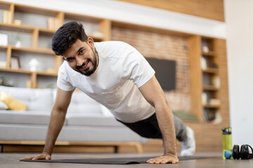 Attractive arabian man standing in plank position on yoga mat at living room. Young guy in sportswear smiling and looking at camera while balancing on straight hands.