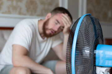 Young bearded man using electric fan at home, sitting on couch cooling off during hot weather, suffering from heat, high temperature - obrazy, fototapety, plakaty