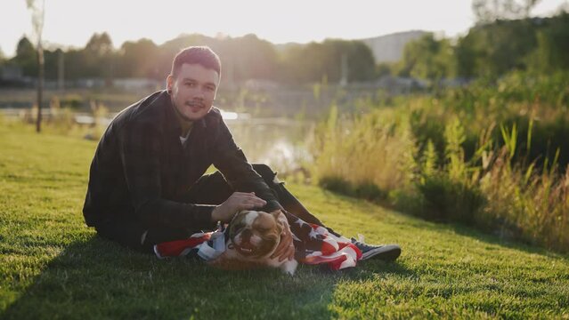Portrait of the handsome smiled stylish man sitting on the grass with mini bull terrier is wrapped in the usa flag, sitting near her in the park. Patriotic dog is wrapped in the american flag