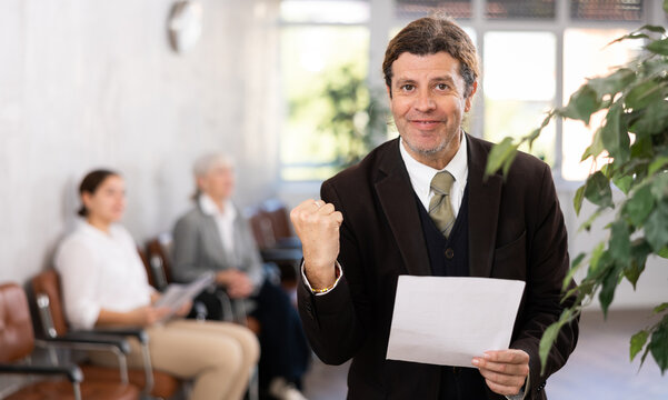 Portrait of happy satisfied adult man standing in office with papers in hands, making triumphant woohoo gesture