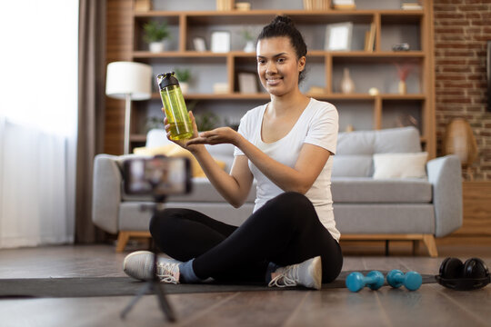 Sporty african american woman sitting in front of digital device on tripod and pointing at sports bottle. Focused fitness instructor explaining importance of drinking fluid during exercise at home. - Powered by Adobe