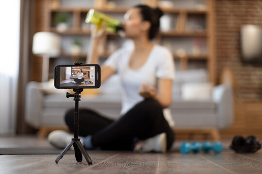 Modern smartphone on tripod recording video of young multiracial lady in athletic wear drinking from sports bottle on camera. Media influencer shooting video for her sports vlog followers indoors.