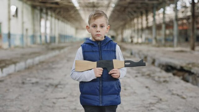 Portrait of the little boy holds a toy gun against a background of destroyed plant looking at camera. War. Male child playing in war