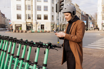 Young man wear helmet and unlocks an electric scooter with his mobile phone. Protective gear in...