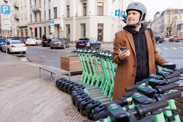 Young man wear helmet and unlocks an electric scooter with his mobile phone. Protective gear in urban city. New way of mobility. Green traffic. Sustainable transportation and climate neutral cities. © Girts