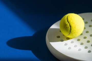White professional paddle tennis racket  and yellow ball with natural lighting on blue background....