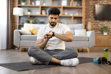 Attractive indian male in athletic wear checking workout achievements on sports watch while sitting on yoga mat in living room. Young adult taking advantages of home training using fitness tracker.