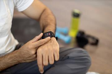 Close-up of young sporty indian man sitting on wooden floor and checking pulse on modern smartwatch...