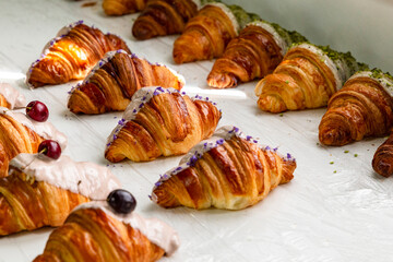 Rows of decorated croissants in a row in a bakery. of cherry-flavored Violet-flavor, cherry-flavor, pistachio-flavored croissants half covered with white chocolate