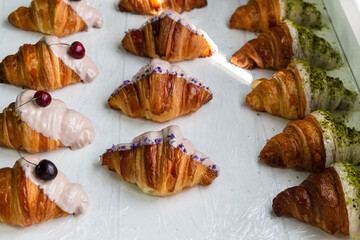 Rows of decorated croissants in a row in a bakery. of cherry-flavored Violet-flavor, cherry-flavor, pistachio-flavored croissants half covered with white chocolate