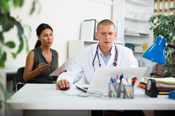 Focused doctor sitting at table with laptop, consulting female patient and writing prescription at clinic..