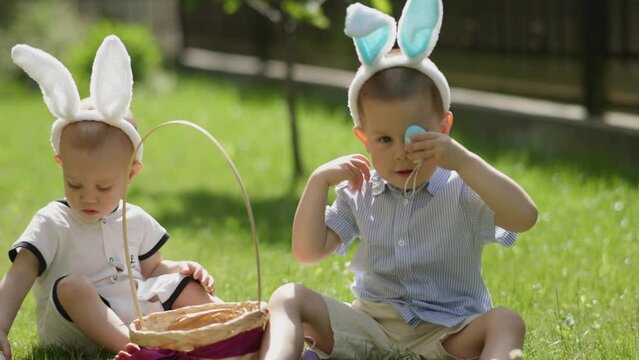 two little Easter children wear bunny ears have Easter picnic on grass outdoors. baby boys siblings have fun playing Easter eggs hide face cover eyes colorful egg traditional Easter holiday in park