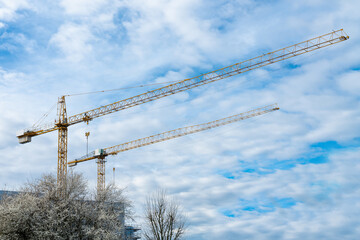 Two yellow construction cranes against a blue sky with light white clouds.
