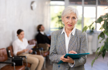 Friendly elderly woman with folder of documents stands in office