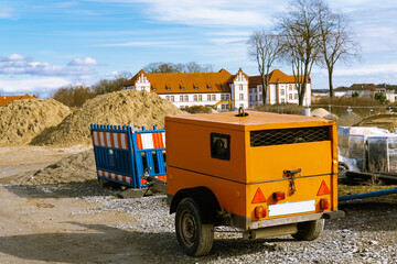 Orange construction generator at a construction site. With heaps of sand and plastic fencing.