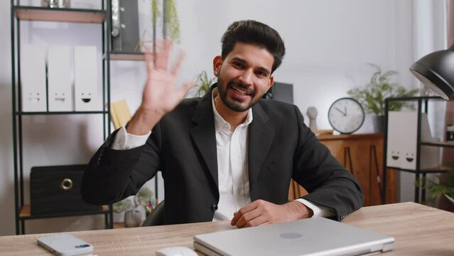Indian business man working on laptop computer smiling friendly at camera and waving hands gesturing hello, hi, greeting or goodbye, welcoming with hospitable expression at home office workplace desk