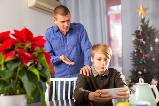 Portrait of teenage boy sitting at table at home interior with smartphone and playing game, father trying to speak with him during Christmas - Powered by Adobe