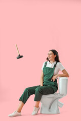 Female plumber sitting on toilet bowl and flying plunger against pink background