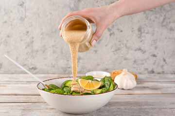 Woman adding tahini into bowl of salad on light wooden table