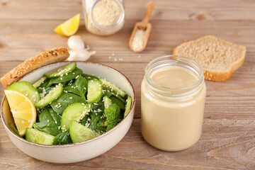 Glass jar of tasty tahini and bowl with salad on wooden table