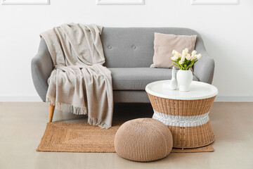 Fototapeta na wymiar Cozy grey sofa with cushion, vase of tulip flowers on table and pouf near white wall