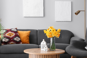 Porcelain houses and vase with narcissus flowers on coffee table near sofa in living room