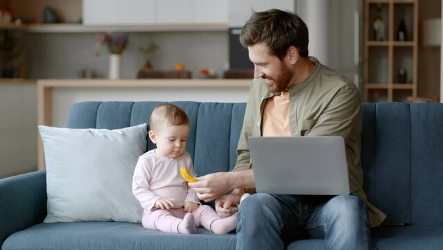 Young positive man independent contractor playing with his baby daughter and typing on laptop, tracking shot, free space