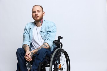 A man in a wheelchair looks thoughtfully at the camera, copy space, with tattoos on his arms sits on a gray studio background, the concept of health a person with disabilities, a real person
