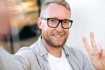 Obraz premium Selfie photo of a positive attractive confident caucasian man with glasses, wearing a casual shirt, standing outdoors, takes a selfie photo on a smart phone, recording online stream, smiling at camera