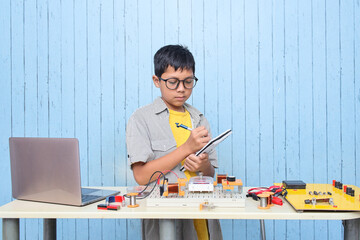 Smart looking Asian boy taking notes while working with circuits, wires, computer, motor on his project. Science, Technology, Engineering and Mathematics (STEM) education concept.