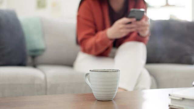Coffee, relax and table with a woman in the background of her home living room typing a text message. Tea, cup and beverage with a mug on a wooden surface in the room of a house interior closeup