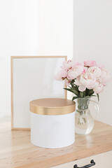 Vertical frame and gift box mockup on a wooden table in the kitchen. Glass jug with a bouquet of pink peonies. Scandinavian style interior.