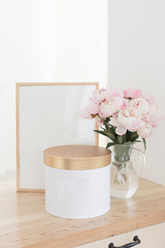Vertical frame and gift box mockup on a wooden table in the kitchen. Glass jug with a bouquet of pink peonies. Scandinavian style interior.