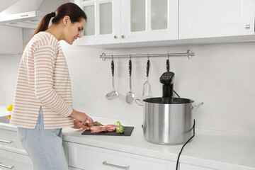 Woman cutting meat near pot with sous vide cooker in kitchen. Thermal immersion circulator