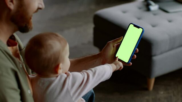Young man web surfing on smartphone with green chroma key screen, curious little baby trying to touch gadget, free space