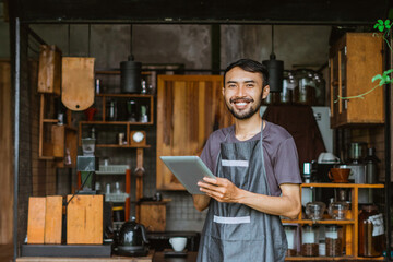 male barista in apron standing in front of the bar desk while using the digital tablet