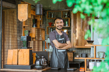 male barista in apron standing with handcrossed inside the bar desk and smiling