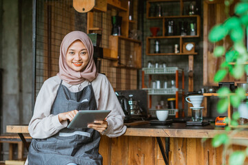 Fototapeta na wymiar muslim female barista smiling while using the digital tablet in front of the bar desk