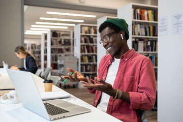 Pleased satisfied African American guy student or freelancer gestures with hands in conversation using laptop for video conference. Smiling black man sitting at library using video call for education.