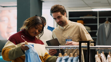 Retail employee helping client to choose modern clothes, woman looking to buy trendy brands from shopping center. Customer talking to assistant about fashion merchandise, new collection.