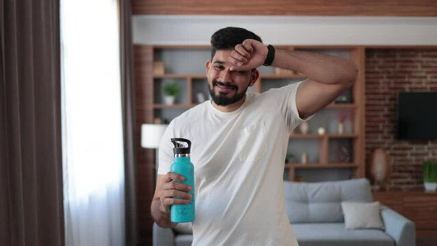 Tired indian man in sport clothes wiping sweat and drinking water during morning exercise. Active young guy having regular workout during free time at home.