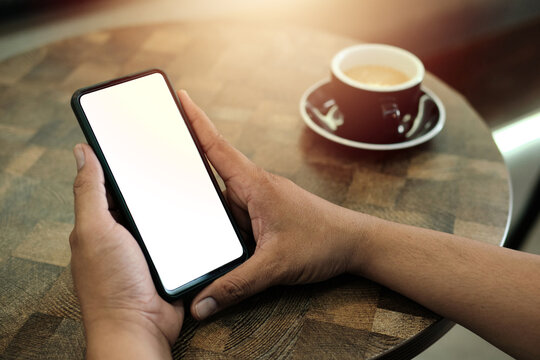 Mockup image of a man holding mobile phone with blank white screen with coffee cup on wooden table
