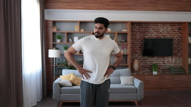 Arabian bearded man dressed in sport clothes holding hands on hips and bending body to sides. Sporty young athlete spending free time for domestic workout.