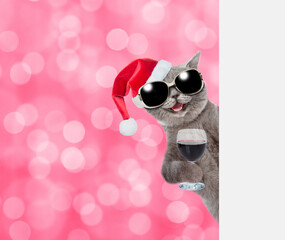 Happy cat wearing sunglasses andred santa hat holds glass of red wine and looks from  behind empty white banner. Shade trendy color of the year 2023 - Viva Magenta background. Empty space for text