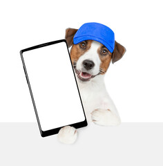 Jack russell terrier puppy wearing blue cap holds big smartphone with white blank screen in it paw above empty white banner. isolated on white background